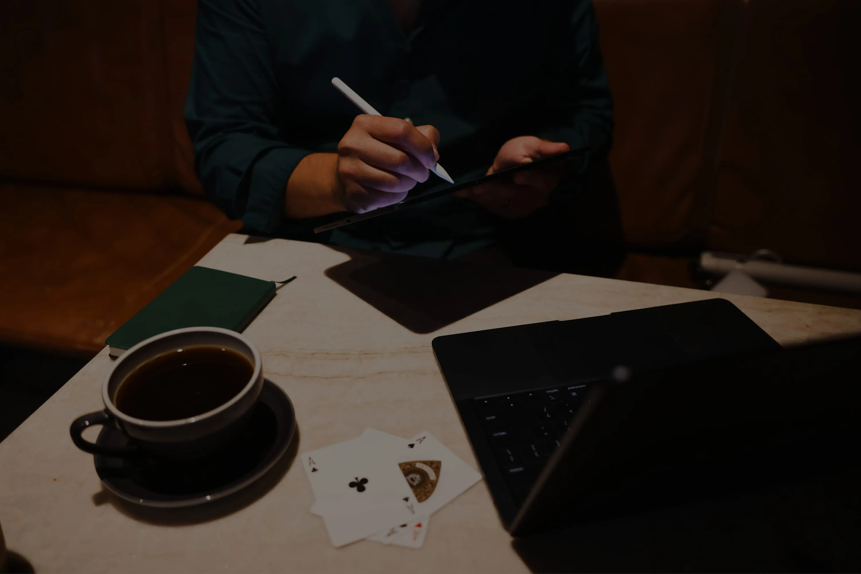 Mauri sketching on a tablet with a stylus at a café table, with a cup of coffee, a green notebook, playing cards and a laptop, in low light