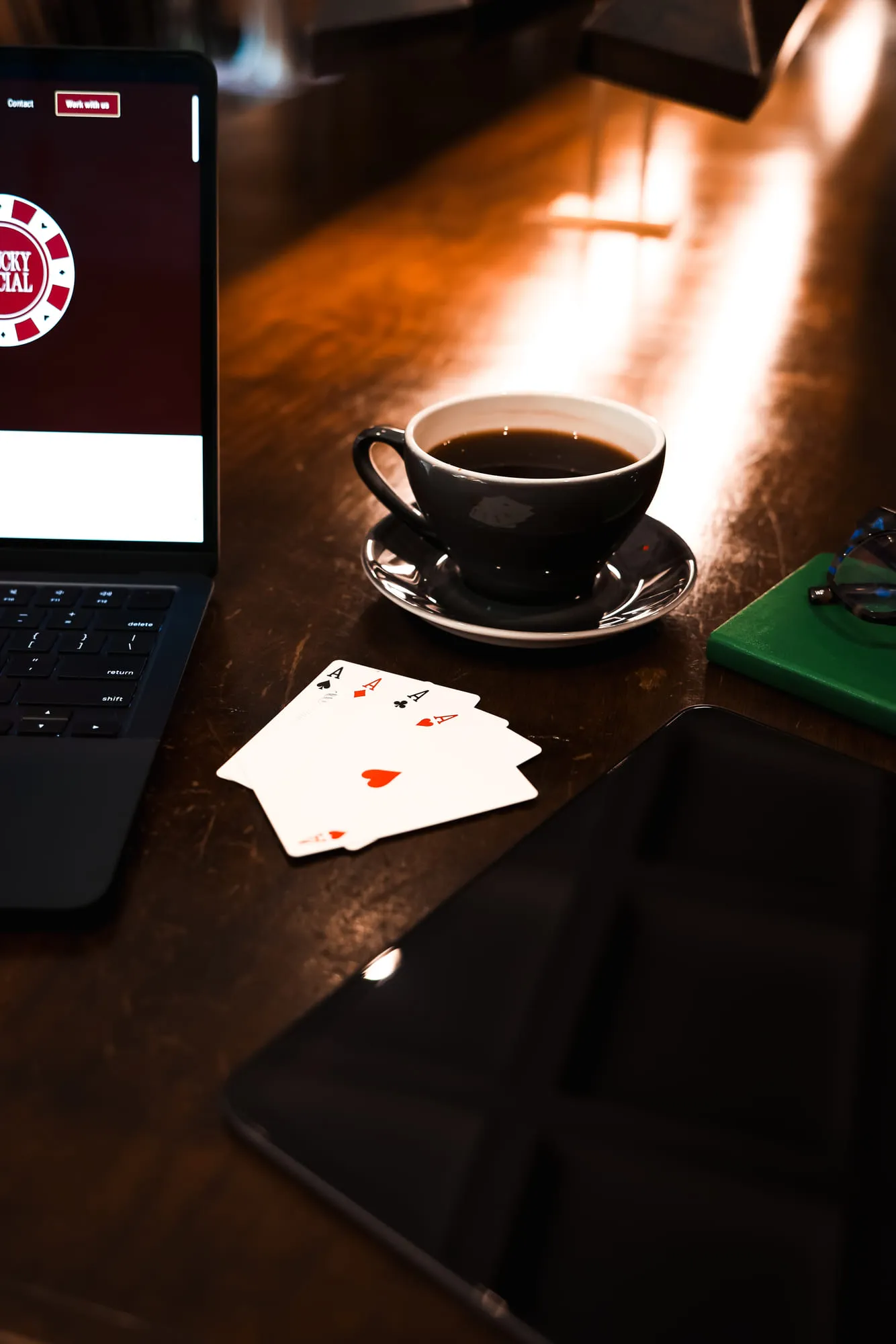 Four aces, a cup of coffee, a tablet and a laptop showing the Lucky Social website on a wood table